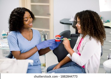 Teeth Hygiene. Cute Happy Child Girl With Red Curly Hair, Having Fun With Her Mom At Dentist Office, While Trying To Brush Teeth On Jaw Model Under The Supervision Of Female Black Dentist