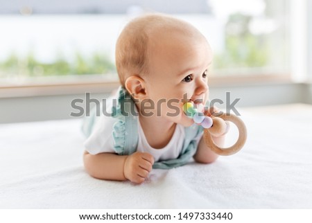 teething, babyhood and people concept - sweet little baby girl lying on white blanket and chewing wooden rattle