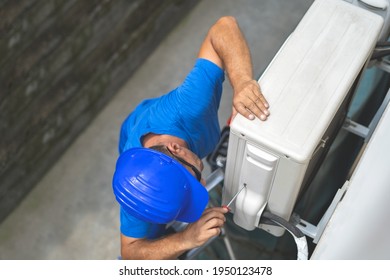 
Tehnician In A Blue Helmet On His Head Stands On The Ladder And Looks At The Screwdriver That Screws The Screw In The Air Conditioning Compressor