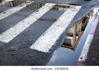 A Tel Aviv building reflecting in a puddle that's between the curb and the crosswalk zebra stripes. Winter.