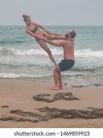 Tel Aviv - June 26th 2019: Muscular man and fit young woman training together in Acro Yoga. Morning sun lighting the couple and the mediterranean beach background.
