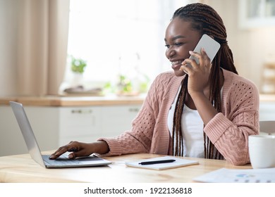 Telecommuting Concept. Black Woman Using Cellphone And Working On Laptop In Kitchen. Smiling African American Freelancer Lady Talking On Mobile Phone And Typing On Computer Keyboard, Having Remote Job