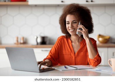 Telecommuting Concept. Black Woman Working On Laptop Computer And Using Cellphone In Kitchen, Smiling African American Female Freelancer Talking On Mobile Phone, Enjoying Remote Job, Copy Space - Powered by Shutterstock