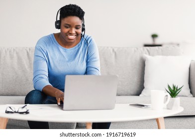 Telecommuting Concept. Smiling Curvy African American Woman Sitting On The Sofa And Working Online On Laptop, Wearing Headset. Young Black Female Using Computer At Home Office, Typing On Keyboard