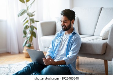 Telecommuting Concept. Young Eastern Guy Freelancer Working On Laptop While Sitting On Floor In Cozy Living Room At Home, Smiling Millennial Arab Man Enjoying Remote Job, Free Space