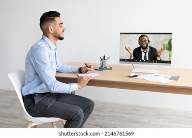 Telecommuting And Distance Education Concept. Smiling Young Arab Manager Making Video Call With Black Man Using Laptop At Office, Having Web Conference Looking At Computer Screen, Writing In Notebook