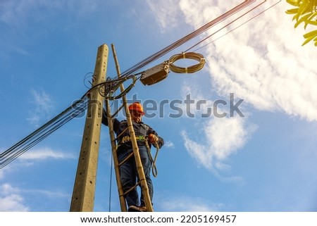 A telecoms worker is shown working from a utility pole ladder while wearing high visibility personal safety clothing, PPE, and a hard hat.	