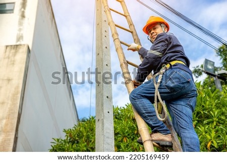 A telecoms worker is shown working from a utility pole ladder while wearing high visibility personal safety clothing, PPE, and a hard hat.	