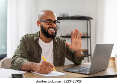 Teleconference and telecommuting. Happy latin man sitting at desk, having video call on laptop, waving to webcamera. Positive male wearing earphones at virtual meeting - Shutterstock ID 2223425929