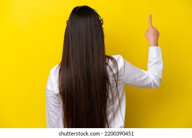 Telemarketer Chinese Woman Working With A Headset Isolated On Yellow Background Pointing Back With The Index Finger