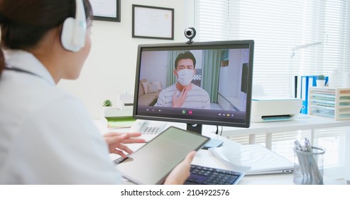 Telemedicine Concept - Asian Female Doctor Talking To Patient Online Through Webcam With Headsets And Young Male Patient Wearing Face Mask Is Talking About His Sick Symptom From Home