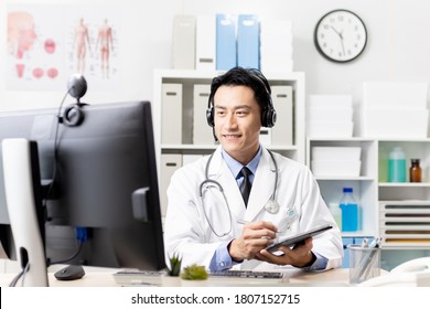 Telemedicine Concept - Asian Male Doctor Wear Headset Talking To Patient And Writing Notes On Digital Tablet Online