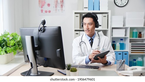 Telemedicine Concept - Asian Male Doctor Wear Headset Talking To Patient By Webcam And Writing Notes On Digital Tablet