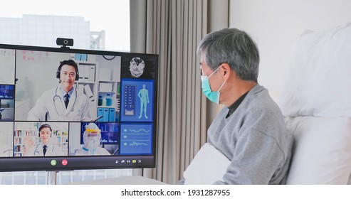 Telemedicine Concept - Asian Senior Man Patient Lying On Bed Has Video Call With Doctor Team By Big Screen TV In A Hospital Ward Or At Home