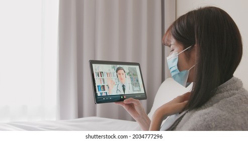 Telemedicine Concept - Asian Woman Patient Wearing Face Mask Is Talking About Her Sick Symptom To Male Doctor By Digital Tablet On Bed