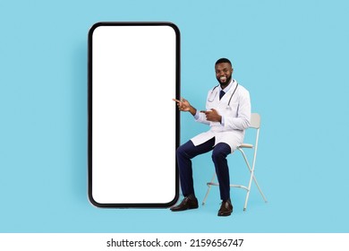 Telemedicine Concept. Friendly Black Doctor Sitting On Chair Near Big Blank Smartphone And Pointing On White Screen, Happy African Male Physician Recommending App For Online Medical Services, Mockup