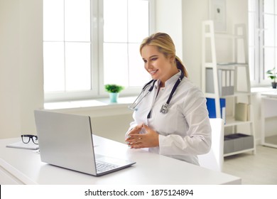 Telemedicine Concept. Friendly Smiling Female Doctor Sitting At Desk In Hospital Office Using Laptop Computer For Video Call With Patient, Giving Health Consultation, Communicating With Client Online