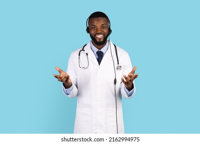 Telemedicine Concept. Positive Black Doctor Wearing Unform And Headset Talking At Camera, Friendly African American Physician Consulting Patient While Standing Isolated On Blue Background, Copy Space