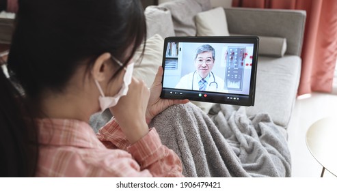 Telemedicine Concept - Rear View Of Asian Female Has Video Chat With Elderly Male Doctor By Digital Tablet