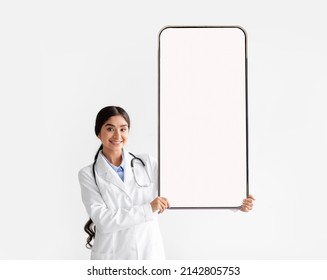 Telemedicine. Happy Female Physician In Uniform And Stethoscope Holding Big Giant Cell Phone Screen In Hands, White Studio Background. Young Doctor Presenting Blank Smartphone Display For Mock Up