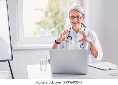 Telemedicine. Middle age female doctor making video call or live video on social network with patient or student while sitting on modern office in hospital - Powered by Shutterstock