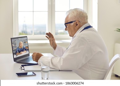 Telemedicine Virtual Visit To Hospital Concept. Doctor Sitting At Desk In EHealth Service Office, Waving Hand At Laptop Computer, Saying Hello To Patient. Senior Man Video Calling His Wife From Work