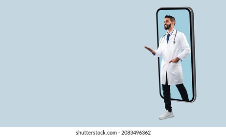 Telemedicine. Young Happy Male Middle Eastern Doctor In White Uniform With Stethoscope Coming Out Big Cell Phone Screen, Stepping Walking Off Gadget Monitor, Showing Free Copy Space, Panorama Banner - Powered by Shutterstock