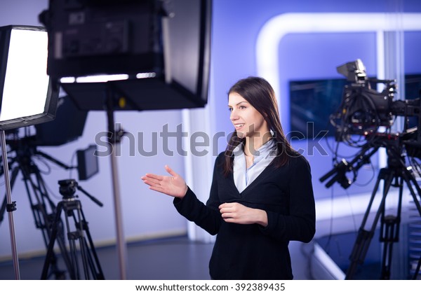 Television presenter recording in television\
news studio.Female journalist anchor presenting business\
report.News camera,light equipment behind the scenes.Talking at\
camera to the TV\
audience.