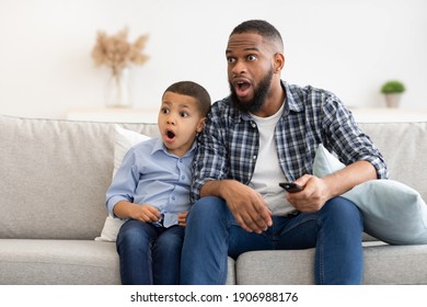 Television Programming Problem. Shocked African Father And Son Watching Questionable Content On TV Together Sitting On Couch With Opened Mouth At Home. Parental Control Concept