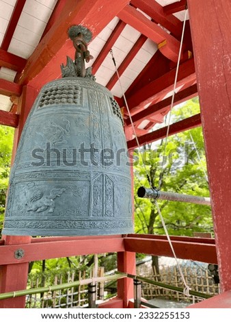 A temple bell hanging from the ceiling of an outdoor red color structure in trees background