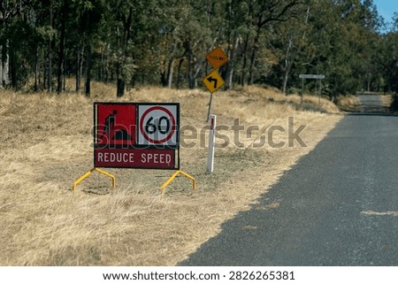 Temporary roadwork sign warns drivers to reduce speed to 60 near a floodway and bend on a quiet rural road through dry woodland.