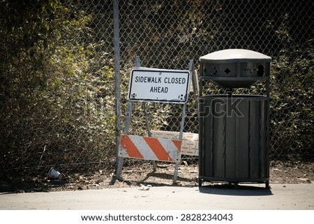 A temporary sign indicates a sidewalk closure ahead, next to a metal trash receptacle and a chain-link fence