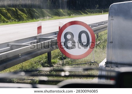 Temporary speed limit marks roadside. Large sign displays eighty restriction. Highway barrier runs beside asphalt. Road safety controls vehicle speed. Traffic regulation forms transport background.
