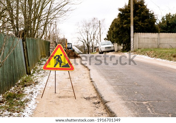 A temporary yellow asphalt road sign has been\
installed on the side of the road: repair work. Traffic rules\
triangle sign. A man with a shovel. Bad quality countryside way.\
Danger. Driver attention.