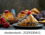 A tempting chocolate croissant topped with fresh strawberries sits invitingly on a wooden table, surrounded by more strawberries, evoking a sense of indulgence and a perfect breakfast treat.