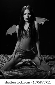 Temptress concept. Demon sexy girl sit on silk sheets. Woman on passionate face play role game. Girl sexy demon with wings, devil full of desire. Lady sexi dressed as demon, devil, black background.