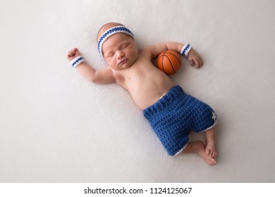 Ten day old newborn baby boy wearing basketball shorts and sweat bands. He is sleeping on his back and has a tiny basketball nestled in his arm.