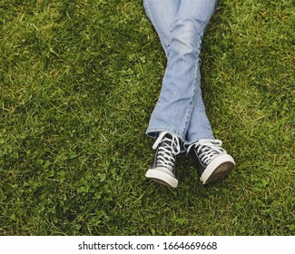 A ten year old girl lying on the grass. Cropped view of her lower legs. Wearing sneakers and faded blue jeans. Legs crossed at the ankles.