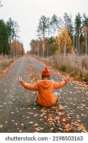 Ten Years Girl In Orange Coat Sits In Yoga Pose On The Road In Fall Forest. Dry Grass And Golden Trees. Autumn Vibes, Outdoor Lifestyle. Deep In Thoughts, Daydreaming And Mental Health