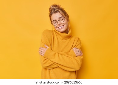 Tender Adorable Woman With Combed Fair Hair Embraces Herself Smiles Gently Keeps Eyes Closed Enjoys Softness Of Cashmere Jumper Feels Glad Isolated Over Vivid Yellow Background. Self Love Concept