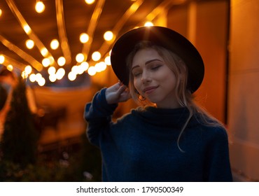 Tender Beautiful Adorable Woman Female Portrait With Smile And Black Hat On Night City Street Restaurant Patio Background View Yellow Illumination Bokeh Glare Of Light Effect 