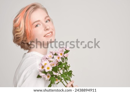 A tender blonde girl with a short haircut poses with pink flowers on a white studio background. Delicate feminine casual spring-summer look. Beauty portrait. Copy space.