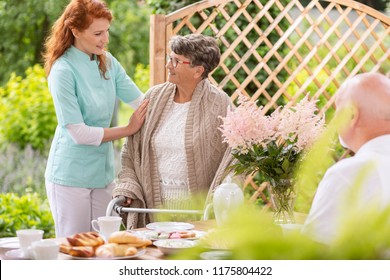 A Tender Caretaker Assisting An Elderly Woman With A Walker During An Afternoon Snack Time On A Patio In The Garden Of A Private Nursing Home.