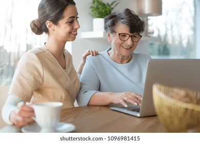 Tender Caretaker Closing The Generation Gap And Teaching A Smiling Senior Woman The Use Of Internet On A Laptop While Sitting By A Table In A Bright Room