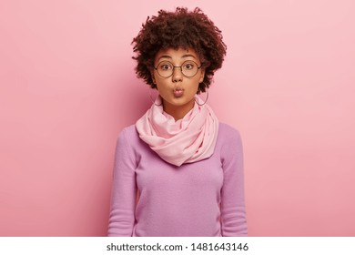 Tender feminine woman keeps lips folded, wants to kiss boyfriend, wears spectacles, hoop earrings, silk scarf around neck, looks straightly at camera, isolated on rosy wall. Human facial expressions