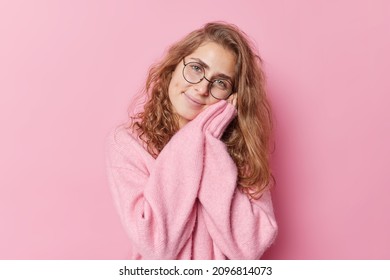 Tender Gentle Young European Woman Keeps Hands Near Face Smiles Happily Wears Round Spectacles And Cashmere Jumper Isolated Over Pink Background. Smiling Long Haired Female Model Gazes At Camera