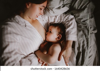 Tender mom and toddler baby, mom breastfeeds the baby in the bedroom in a real interior. The concept of breastfeeding after a year, toning and dark style, top view