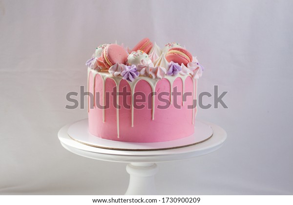Tender pink cake decorated with melted white\
chocolate, macaroons, meringues, cake pops and candies on white\
cakestand. Plain\
background.