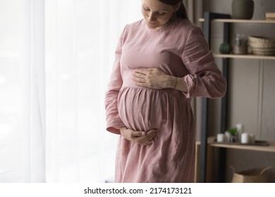 Tender Pregnant Expectant Mother In Long Elegant Dress Standing In Living Room Touch Her Belly Feels Love To Unborn Baby Standing Alone At Home. Medical Insurance Cover, Maternity, Motherhood Concept