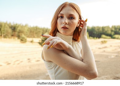 Tender redhead young woman adjusting long hair while standing on sandy beach in summer season and looking at camera  - Powered by Shutterstock